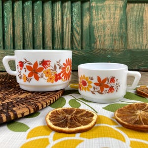 Op de afbeelding: Twee witte keramische mokken met oranje, gele en rode bloemmotieven. De mokken zijn gestapeld en staan op een geweven onderzetter. Gedroogde sinaasappelschijfjes en een patroonkleed.