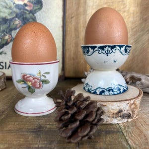 Peut inclure: Deux œufs bruns dans des coquetiers décoratifs. L'une des tasses a un motif floral, l'autre un motif bleu et blanc. Un cône de pin est devant les tasses. Les tasses sont sur une surface en bois.