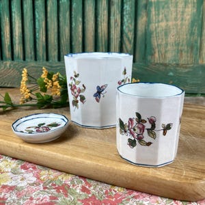 Peut inclure: Ensemble de trois pièces en céramique : deux tasses octogonales et une petite soucoupe. Les tasses et la soucoupe sont blanches avec un motif floral et papillon rose, rouge et bleu. Les bords sont bordés de bleu. Les articles sont sur une planche de bois.
