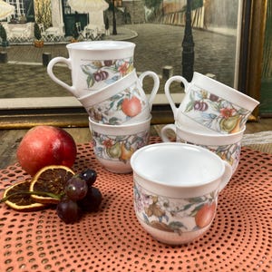 Puede incluir: Un conjunto de tazas de café de cerámica blanca con asas, decoradas con un diseño de frutas y flores. Un melocotón, rodajas de naranja secas y uvas están dispuestas cerca. Las tazas están apiladas sobre una superficie rosa.
