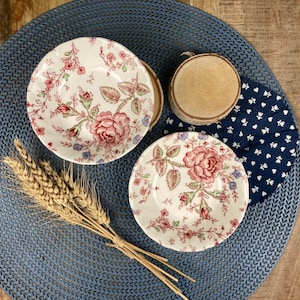 Op de afbeelding: Twee witte keramische kommen met een bloemenpatroon in tinten roze, rood en groen. De kommen staan op een blauwe geweven placemat met een witte stoffen servet met een donkerblauw en wit patroon.