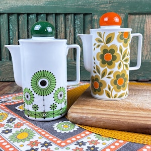 Peut inclure: Deux cafetières en céramique blanche avec poignées et becs verseurs. La cafetière de gauche présente un motif floral circulaire vert et un bouton de couvercle vert. Celle de droite arbore un imprimé floral orange et vert olive.