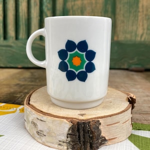 Op de afbeelding: Witte keramische mok met een blauw, groen en oranje bloemmotief. De mok heeft een oor en staat op een houten onderzetter. De mok is een standaardmaat voor koffie of thee.