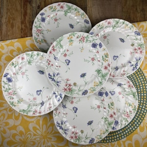 Puede incluir: Juego de seis platos de cena de cerámica blanca con un patrón floral en tonos de azul, rosa y verde. Los platos tienen un borde festoneado y están apilados.