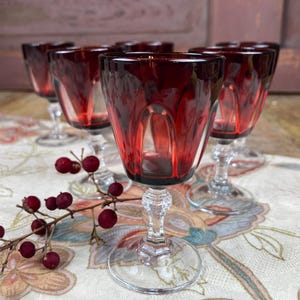 Op de afbeelding: Een set van zes robijnrode wijnglazen met heldere, sierlijke stelen. De glazen staan op een tafelkleed met bloemenpatroon, met een takje rode bessen op de voorgrond. De glazen hebben een klassiek, elegant ontwerp.