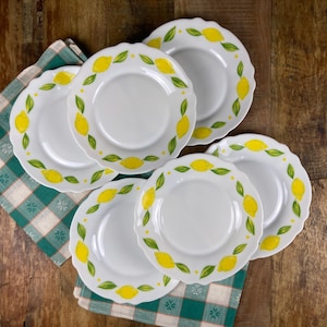 Könnte beinhalten: Ein Set aus sechs weißen Keramikplatten mit gewelltem Rand. Jede Platte ist mit einem Rand aus gelben Zitronen und grünen Blättern verziert. Die Teller sind auf einem grün-weiß karierten Tuch auf einer Holzoberfläche angeordnet.