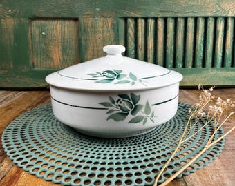 May include: White ceramic serving dish with a lid, featuring a green floral design. The dish has a round shape and is placed on a green, circular, perforated mat. The background includes a wooden surface and a green, slatted door.