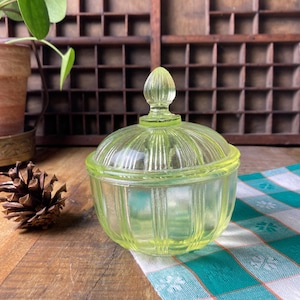 Könnte beinhalten: Ein Vintage-Glasbehälter mit Deckel in Hellgrün mit vertikalen Rippendetails. Der Behälter hat eine abgerundete Form und einen dekorativen Knauf auf dem Deckel. Der Behälter wird auf einer Holzoberfläche mit einem grün-weiß karierten Tuch präsentiert.