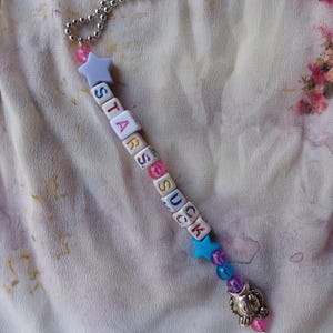 May include: A colorful beaded keychain with a star charm and a silver charm in the shape of a cat's head. The beads spell out the phrase "STARS SUCK".