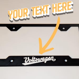 Può includere: Cornice nera per targa con la scritta "Volkswagen" in corsivo bianco. La cornice è rettangolare con angoli arrotondati e fori preforati. Il testo "YOUR TEXT HERE" è sopra la cornice.