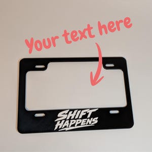 May include: A black rectangular licence plate frame with four mounting slots. The bottom centre features the white text SHIFT HAPPENS in a stylised font. Above the frame, the pink text Your text here appears with a pink arrow pointing down.