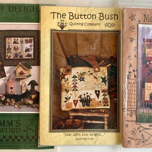 May include: Three quilting pattern books in green, brown and cream colours. The first book is titled "Garden of Delight" and features a quilt design with a birdhouse and a garden scene. The second book is titled "The Button Bush Quilting Company" and features a quilt design of a tote bag with a star and heart pattern. The third book is titled "Mumm's Quilts" and features a quilt design with a chicken and a farmhouse scene.