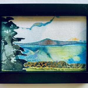 Peut inclure: Une œuvre d'art textile encadrée représentant un paysage avec la silhouette sombre d'un arbre sur la gauche, une étendue d'eau bleue et verte, une chaîne de montagnes lointaine et un premier plan texturé avec des coutures jaune-vert.