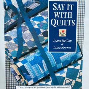 Könnte beinhalten: Ein Buch mit dem Titel "Say It With Quilts" mit blauem Einband, das einen Stapel blau-weißer Quilts zeigt. Das Buch hat ein blaues Band und den Text "23 New Quilts from the Authors of Quilts, Quilts, and More Quilts!"