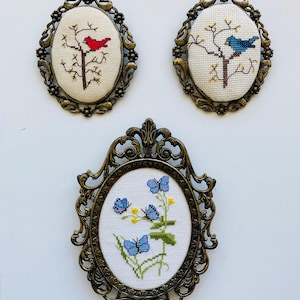 May include: Three oval wall hangings featuring cross-stitch embroidery in ornate bronze-toned metal frames. Two pieces depict birds on branches, one red and one blue, while the third piece displays blue butterflies and yellow flowers on white fabric.