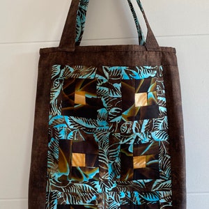 May include: A brown canvas tote bag with blue and gold patterned fabric squares. The bag has two brown straps.