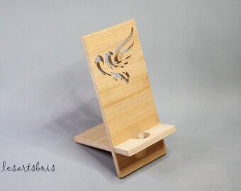 Puede incluir: Un soporte de teléfono de madera con un diseño de pájaro recortado. El soporte está hecho de madera de bambú marrón claro y tiene un pequeño borde para sujetar un teléfono.