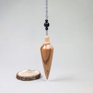 May include: A wooden pendulum with a pointed end, suspended from a silver chain with black beads. The pendulum is light brown with a natural wood grain. A small, round wooden slice sits on the surface below.