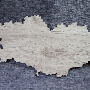 Op de afbeelding: Een houten uitsparing in de vorm van een geografische regio, met een lichte houtnerf afwerking. De randen zijn ongelijkmatig, wat wijst op een ambachtelijke of natuurlijke esthetiek. De uitsparing staat tegen een grijze achtergrond.