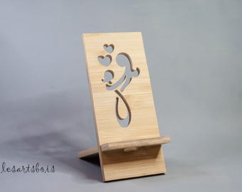 Puede incluir: Un soporte de teléfono de madera con un diseño recortado de una persona sosteniendo un corazón. El soporte está hecho de madera marrón claro y tiene un pequeño estante para sostener un teléfono.
