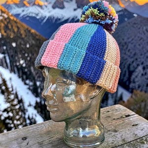 May include: A colorful knit beanie with a striped pattern in pink, blue, and beige, topped with a multi-colored pom-pom. The hat is displayed on a glass head, with a mountain range in the background.