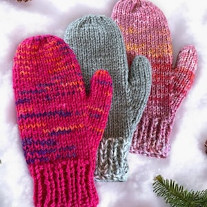 May include: Three pairs of knitted mittens in pink, gray, and a bright pink with orange and purple stripes. The mittens are lying on a white surface with pine branches and pine cones.