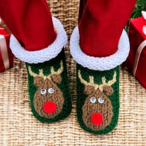 May include: Green knitted Christmas slippers with a reindeer design, featuring a brown face, red nose, and white eyes. The slippers have a white knitted cuff and are worn with red trousers. The image is taken from a low angle.