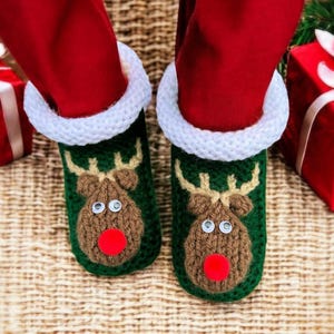 Può includere: Pantofole natalizie verdi lavorate a maglia con un design a renna, con viso marrone, naso rosso e occhi bianchi. Le pantofole hanno un polsino lavorato a maglia bianco e sono indossate con pantaloni rossi. L'immagine è ripresa dal basso.