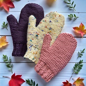 May include: Three knitted mittens in shades of purple, speckled cream, and pink, arranged on a light blue wooden surface with autumn leaves and greenery. The mittens are handmade, showcasing a cozy, textured design, perfect for cold weather.