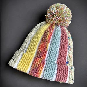 May include: A knit beanie featuring vertical stripes in yellow, orange, light blue, and pink, separated by white bands. The hat has a speckled cream-colored base and is topped with a multicolored pom-pom made of yarn in pastel shades of green, pink, and yellow.