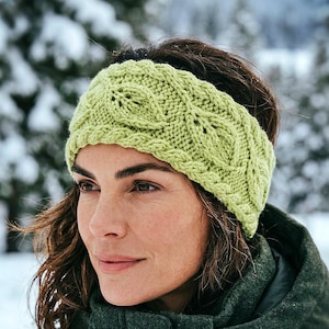 May include: A light green knitted headband with a cable knit design. The headband is worn by a person in a dark green jacket, with a snowy, blurred background of trees.