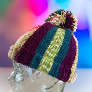 Könnte beinhalten: Eine bunte gestreifte Strickmütze mit einem Bommel oben. Die Mütze ist aus weichem, warmem Garn gefertigt und perfekt, um den Kopf im Winter warm zu halten.