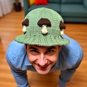 May include: A green knitted bucket hat with brown and white mushroom appliqués. The hat has a wide brim and a ribbed texture. The hat is worn by a person wearing a blue shirt.