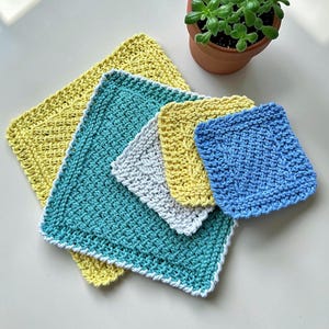 May include: A collection of handmade crochet washcloths in various sizes and colors, including yellow, teal, gray, and blue. The washcloths are arranged on a white surface, with a small potted plant in the background. The crochet stitch creates a textured appearance.