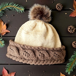 以下が含まれることがあります： クリーム色のニット帽、茶色のケーブル編みのつば、ふわふわの茶色のポンポン。 帽子は、葉、松葉、松ぼっくりなど、秋の葉に囲まれ、暗い木製の表面に置かれています。