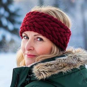May include: A woman wearing a burgundy knitted headband and a dark green winter coat with a fur-lined hood. The headband is a thick, braided style. The background is a snowy winter scene with evergreen trees.