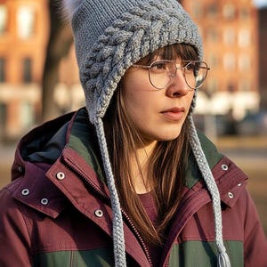 Può includere: Un cappello di lana grigio con un motivo a trecce e un pompon in finta pelliccia bianca. Il cappello ha paraorecchie con nappe. La persona nell'immagine indossa una giacca bordeaux e verde. È un accessorio invernale.