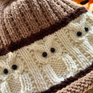 May include: A brown and white knitted hat with a brown faux fur pom pom. The hat has a brown stripe and a white stripe with black buttons.