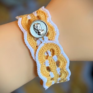 Könnte beinhalten: Ein gelbes und weißes gehäkeltes Armband mit einem silbernen Knopf mit einem erhabenen Design.