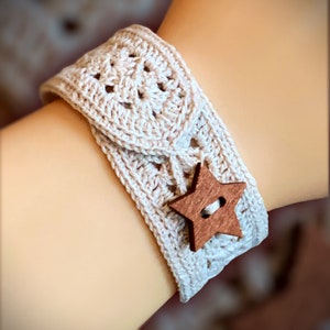 May include: A cream-colored, crocheted bracelet with a star-shaped wooden button closure. The bracelet is approximately 2 inches wide and features a textured pattern. The wooden button is a medium brown color.
