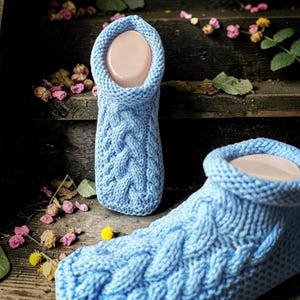Op de afbeelding: Een paar lichtblauwe gebreide pantoffels met een opvallend verticaal kabelpatroon op de bovenzijde en een dikke, omgerolde boord. De pantoffels zijn gemaakt van een getextureerd garen in een consistente lichtblauwe kleur.