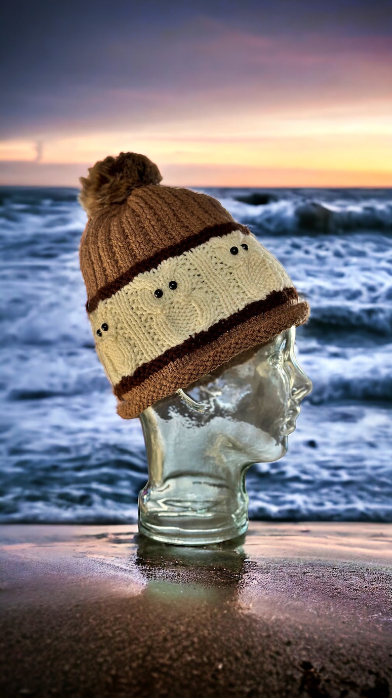May include: A brown and white knit beanie with a pom pom on top. The beanie has black buttons on the front. The beanie is sitting on a glass head.