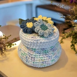 Könnte beinhalten: Ein handgefertigter Häkelbehälter mit Deckel, verziert mit drei gehäkelten Blumen in Blau- und Gelbtönen. Der Behälter ist weiß mit pastellfarbenen Sprenkeln. Die Blumen sind am Deckel befestigt.