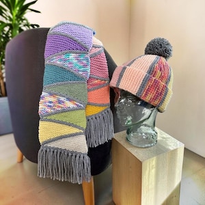 May include: A hand-knitted scarf and matching beanie set featuring a geometric pattern of triangular segments in lavender, teal, olive green, yellow, and pink. The scarf has grey fringe at both ends, and the beanie has a grey pom-pom on top.