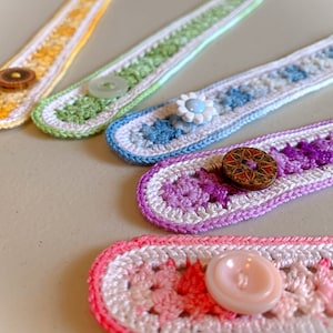 May include: Five crocheted mask extenders with different coloured stripes and buttons. The extenders are made with white and coloured yarn. The buttons are made of wood and plastic.
