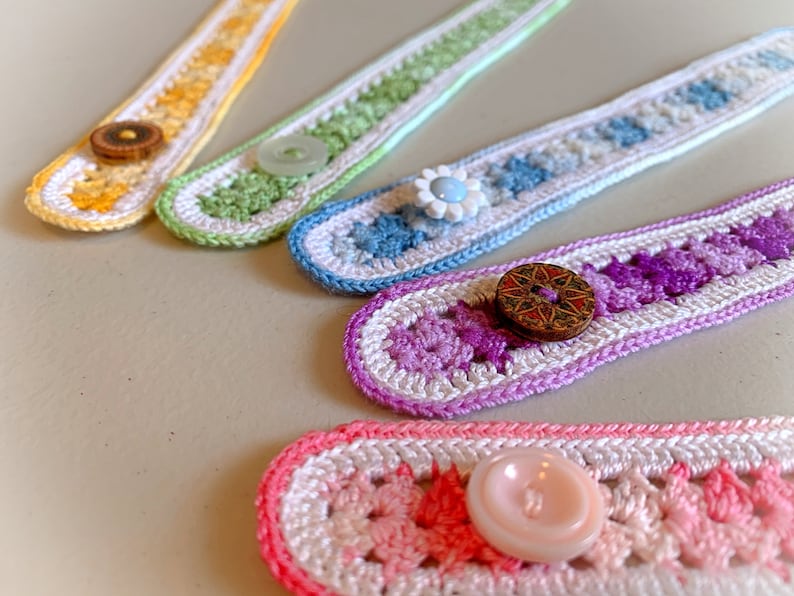 May include: Five crocheted mask extenders with different coloured stripes and buttons. The extenders are made with white and coloured yarn. The buttons are made of wood and plastic.