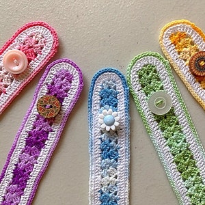 May include: Five colourful crocheted mask extenders with different coloured buttons. The extenders are made with white and coloured yarn in a striped pattern. The buttons are pink, blue, gold, and brown.