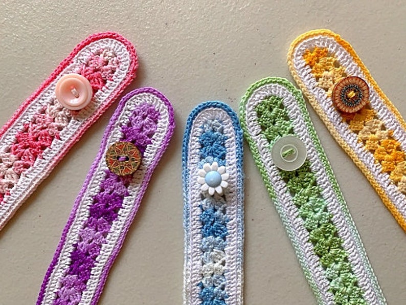 May include: Five colourful crocheted mask extenders with different coloured buttons. The extenders are made with white and coloured yarn in a striped pattern. The buttons are pink, blue, gold, and brown.