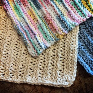 May include: Three knitted dishcloths in different colors and patterns. One is a light beige with a textured pattern, one is a light blue with a textured pattern, and one is a multi-colored striped pattern.