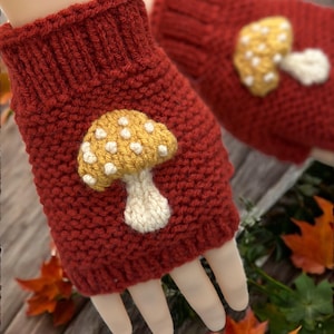 May include: A pair of red knitted fingerless gloves with a yellow and white mushroom applique. The gloves are made of a thick, textured yarn and are designed to keep hands warm while leaving fingers free.